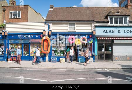 Whitstable England - August 18 2019; People passing Georges Mini Market with beach toys inflatable rings and boat displayed on High Street. Stock Photo