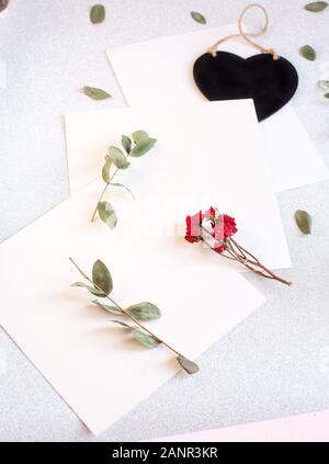 Background with copy space blank on table with black heart, eucalyptus branch, roses flowers and leafs. White paper top view, flat lay, minimal style. Stock Photo