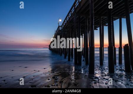 Ventura Pier at sunset, Ventura, California. Rocks and sand in foreground, water receded in low tide. Lamps on pier; colored sky in background. Stock Photo