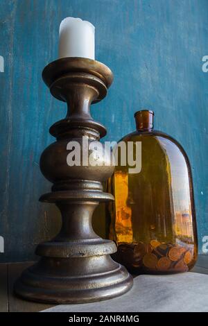 Big brown glass jar with coins and massive candleholder with white candle on it on blue shabby wall background, concept of savings, trading, savings Stock Photo