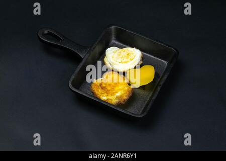 A small black frying pan with quail eggs in the shape of a heart, next to it are bell pepper on the black background. Top views Stock Photo