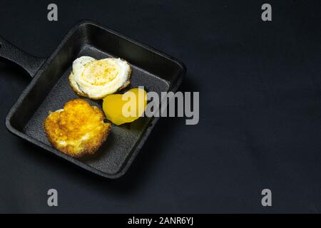 A small black frying pan with quail eggs in the shape of a heart, next to it are bell pepper on the black background. clear space Stock Photo
