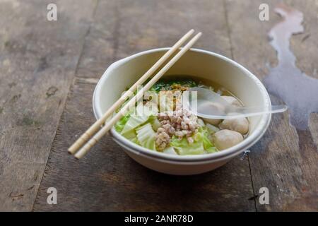 Rice noodle soup with pork balls, minced pork, cabbage and spring onion, serve in white foam bowl and wooden chopstick, on rough wooden. Stock Photo