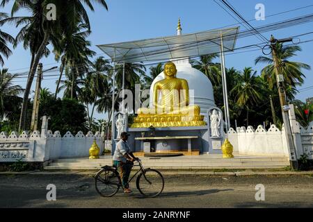 Galle, Sri Lanka - January 2020: A man riding a bicycle passing in front of the Buddha statue on a street in Galle on January 15, 2020 in Galle, Sri L Stock Photo