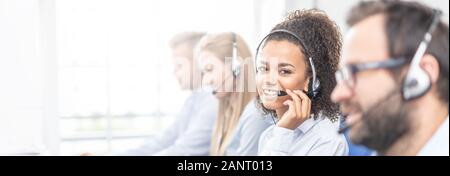 Call center worker accompanied by her team. Smiling customer support operator at work. Young employee working with a headset. Stock Photo