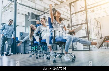 Young cheerful business people dressed in casual clothing are having fun on rowing chairs in a modern office. Happy team concept. Stock Photo