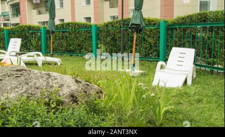 Empty plastic white sun loungers and folded green umbrellas stand on the green grass, in a private area, fenced off by a hedge, rest in a hotel at the Stock Photo