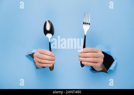 Female hands holding spoon and fork through torn blue paper wall. Gesture of eating dinning. Cooking, healthy homemade food concept. Copy space Stock Photo