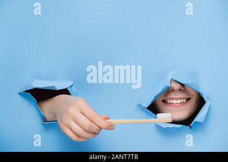 Woman with toothy pleasant smile on face, keeps head and bamboo eco toothbrush through torn hole in blue paper, copy space for your promotion. Dental Stock Photo