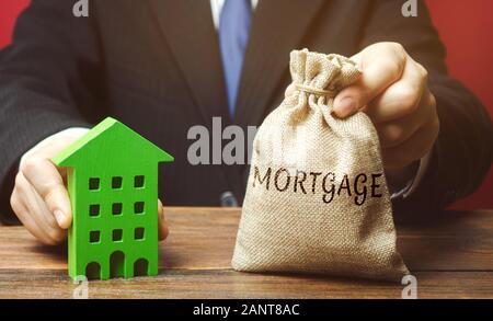 Businessman holds a money bag with the word Mortgage and a wooden house. Real estate concept. Loan for apartment and housing. Credit insurance. Busine Stock Photo