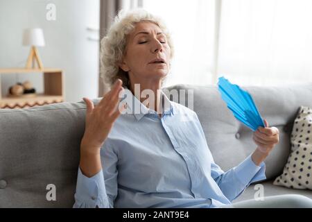Tired older woman waving fan, suffering from heat at home Stock Photo