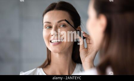 Smiling beautiful woman looking in mirror, applying mascara on lashes Stock Photo