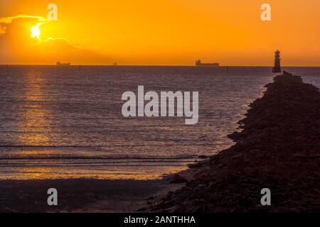 Silhouette of cargo ships at golden hour. Sun, solar disc, shines from behind cloud. Lighthouse and breakwater on Bull Island, Dublin, Ireland Stock Photo