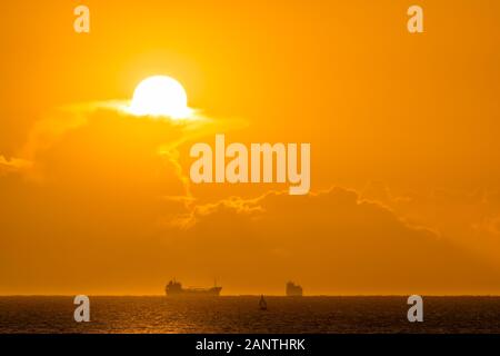 Silhouette of cargo ships on far distance at golden hour. Sun, solar disc, shines from behind cloud. Bull Island, Dublin, Ireland Stock Photo