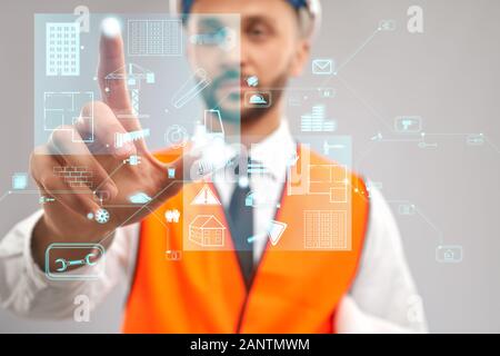 Close up of architect wearing reflective vest and helmet. Selective focus of digital tactile charts screen, man touching virtual icon on projection. Concept of digitalization, construction. Stock Photo