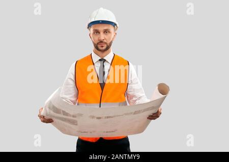 Portrait of young architect in orange reflective vest and helmet holding big map. Engineer crrying construction plan, looking at camera, isolated on grey. Concept of architecture, construction. Stock Photo