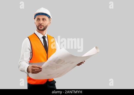 Front view of young bearded architect in orange reflective vest and helmet holding big map. Engineer crrying construction plan, looking up, isolated on grey. Concept of architecture, construction. Stock Photo