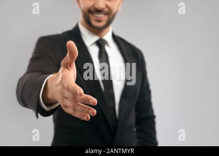 Close up of incognito smiling businessman in black formal suit giving hand for shake. Selective focus of friendly gesture, isolated on grey background. Concept of business, sealing deal. Stock Photo