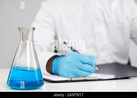 Front crop of incognito chemist in white lab coat and gloves sitting at desk and writing in folder, isolated on grey. Selective focus of flask with blue liquid on table. Concept of chemistry. Stock Photo