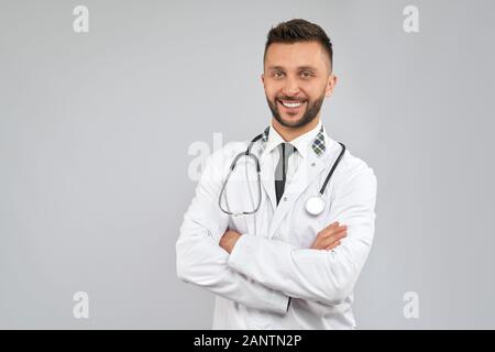 Front view of handsome brunette male doctor wearing lab coat and stethoscope, looking at camera with happy expression. Close up of bearded man with crossed arms isolated on grey. Concept of medicine. Stock Photo