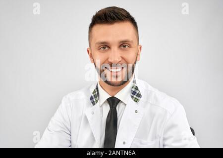 Front view of handsome confident brunette male doctor wearing lab coat and looking at camera with happy expression. Close up of bearded medic sitting, isolated on grey. Concept of medicine.