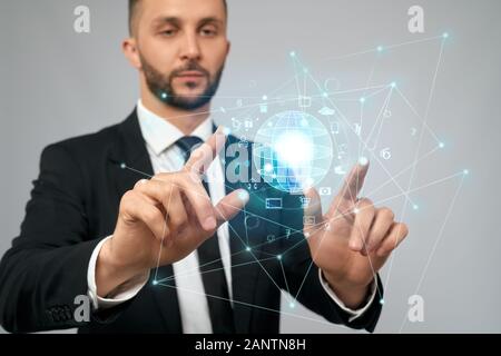 Front view of young serious businessman in black suit in office. Selective focus of digital tactile charts screen, bearded man touching virtual icons. Concept of high technologies, digitalization. Stock Photo