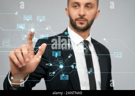 Front view of young serious businessman in black suit in office. Selective focus of digital tactile charts screen, bearded man clicking virtual icon. Concept of high technologies, digitalization. Stock Photo
