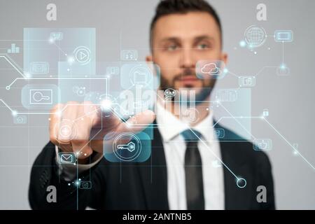 Front view of young serious businessman in black suit in office. Selective focus of digital tactile charts screen, bearded man clicking virtual icon. Concept of high technologies, digitalization. Stock Photo