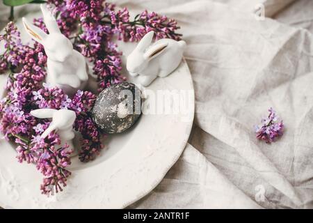 Happy Easter. Stylish Easter white bunnies, modern easter egg on vintage plate and lilac flowers on linen rural fabric. Natural dyed easter egg and sp Stock Photo