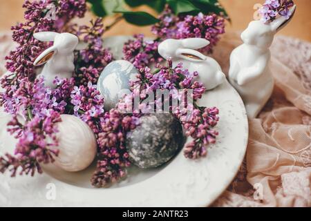Happy Easter. Rustic composition of natural dyed easter eggs, lilac flowers and bunny rabbits on linen fabric on wooden table. White ceramic bunny and Stock Photo