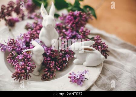 Stylish Easter white bunnies on vintage plate and lilac flowers on rural fabric. Ceramic rabbits decorations and spring flowers. Space for text. Holid Stock Photo