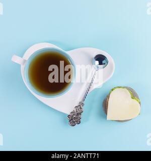 Blue cup of tea and silver spoon on white saucer in heart shape and sandwich with cheese and herbs in heart shape on stone on light blue background. R