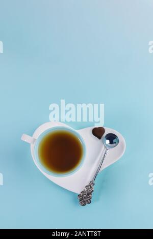Blue cup of tea with silver spoon and one chocolate candy on white saucer in heart shape on light blue background. Romantic breakfast. Valentine's day