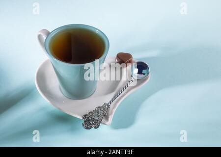 Creative photo with shadows. Romantic breakfast with cup of tea, silver spoon, chocolate candy on saucer in heart shape on blue background. Valentine'