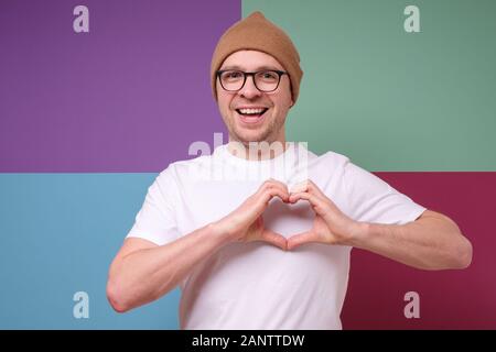 Lovely caucasian young guy smiling cheerfully and making heart gesture over chest, expressing his feelings and tenderness. I am in love with you Stock Photo