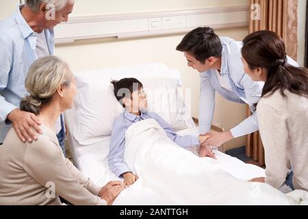 five year old asian child lying in bed in hospital ward gets a visit from parents and grandparents Stock Photo