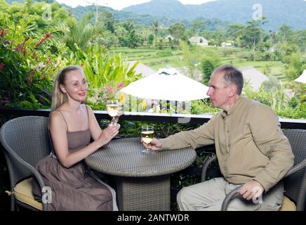 The man and the woman with glasses of wine sit on a balcony overlooking rice terraces and mountains. Stock Photo