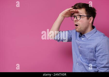 Surprised millennial man in casual clothes and glasses looking aside far away with copyspace and pink wall. Guy pondering considering good sale offer, Stock Photo