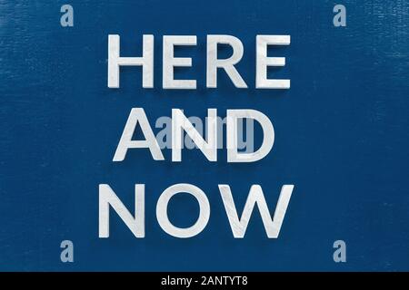 the slogan here and now laid with silver metal letters on classic blue painted board background Stock Photo