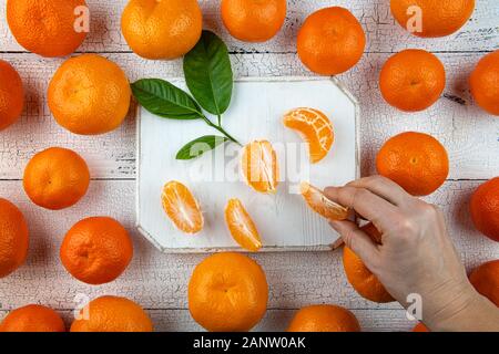 Human hand is taking tangerine segments from square white cutting board surrounded by whole orange tangerines on old wooden table. Fruits have natural Stock Photo