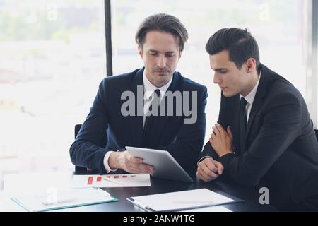 Discussing a new project. Two business men looking at the digital tablet sitting at office desk with financial reports Stock Photo