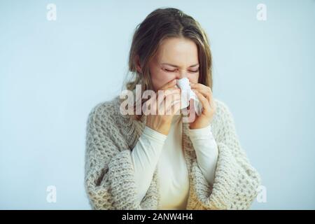 sick elegant woman in roll neck sweater and cardigan with napkin sneezing against winter light blue background. Stock Photo