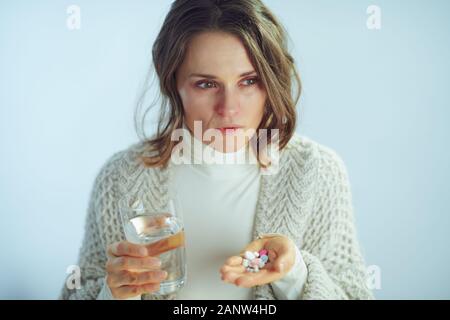 worried sick elegant middle age housewife in roll neck sweater and cardigan with cup of water holding many pills isolated on winter light blue backgro Stock Photo
