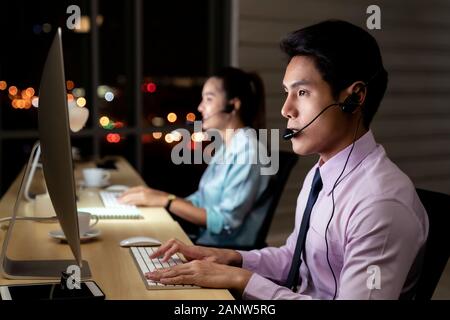 Asian young adult confidence operator man agent with headsets working in a call center at night Environment with his colleague team as customer servic Stock Photo