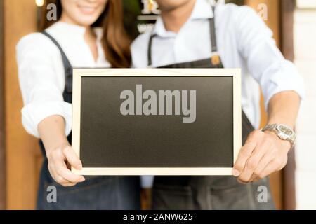 Closeup Asian young adult owners of Small business cafe holding blank chalkboard in front of coffee retail shop. Using as startup SME and consumerism Stock Photo