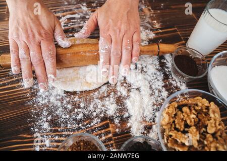 Woman is rolling pin on dough in kitchen, dough recipe ingredients and rolling pin on vintage wood table from above Stock Photo