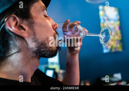 Caucasian man with black hat drinking and tasting glass of red wine with closed eyes, face side selective focus close up view blue interior background Stock Photo