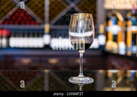 A glass of red wine on black marble countertop selective focus close up view against wine bottles on storage Racks, tasting room blurred background Stock Photo