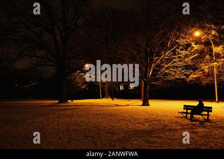 Lady on a bench under the orange glow of street lamps in the local (dog) park on a wintry, snowy evening. Ottawa, Ontario, Canada. Stock Photo