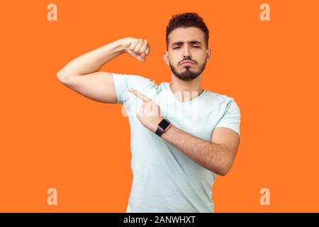 Look at my strength. Portrait of proud handsome brunette man with beard in casual white t-shirt pointing at biceps, feeling confident and powerful. in Stock Photo